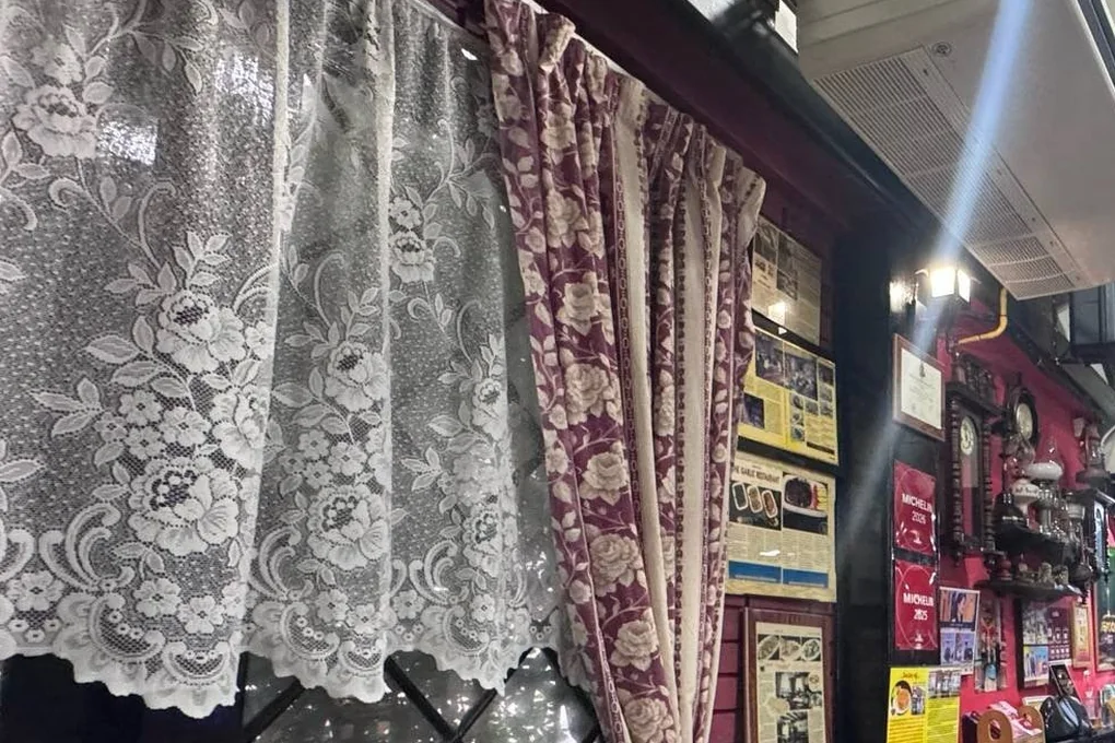 Lace and burgundy curtains at a diamond-paned window, beside a wall of framed cuttings.