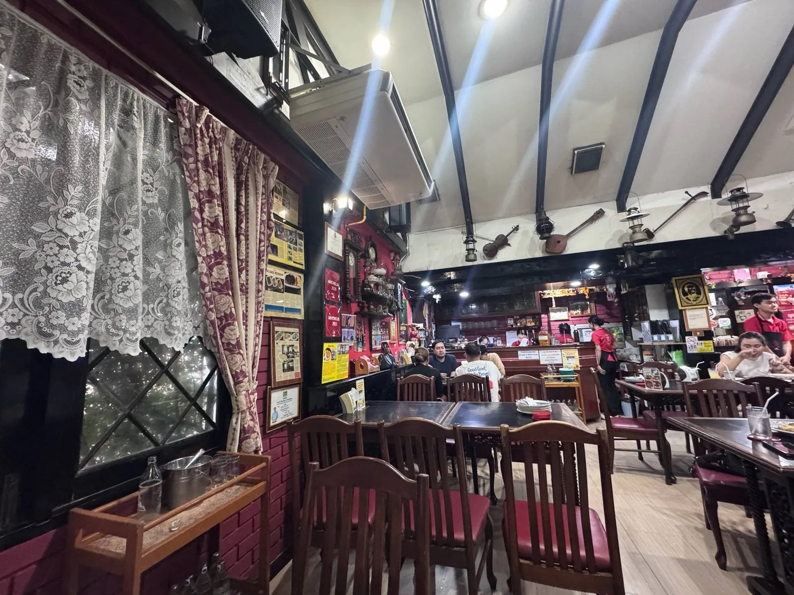 Inside Garlic: oxblood painted brick hung with framed cuttings and certificates, lace curtains, a cream vaulted ceiling on black beams, and brass lanterns.